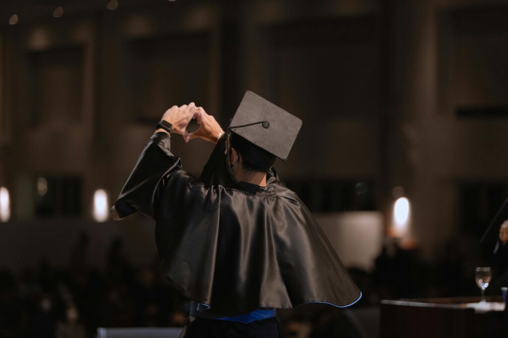 A graduate in cap and gown makes a heart gesture under warm indoor lighting.