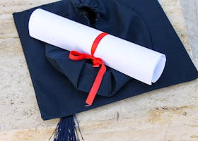 Cost of Attendance Graduation cap with diploma roll tied with red ribbon on concrete surface.