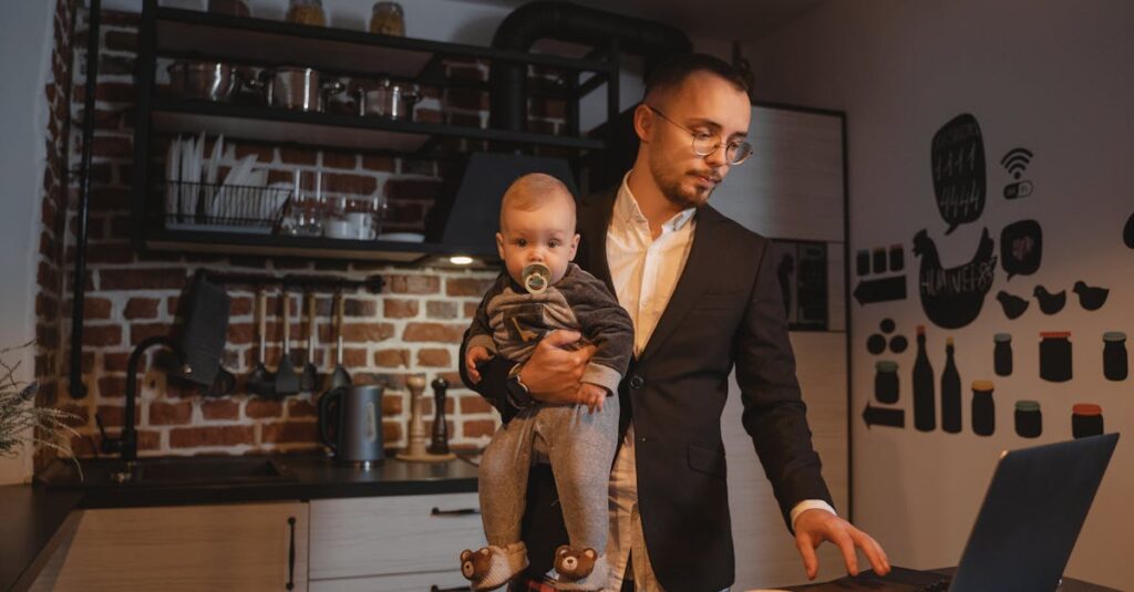 Father working on a laptop while holding his baby in a kitchen setting.