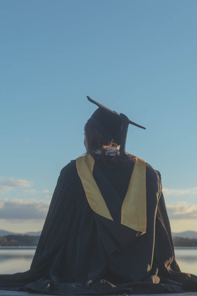 Graduate woman in a cap and gown reflecting by a serene lake at sunset.
