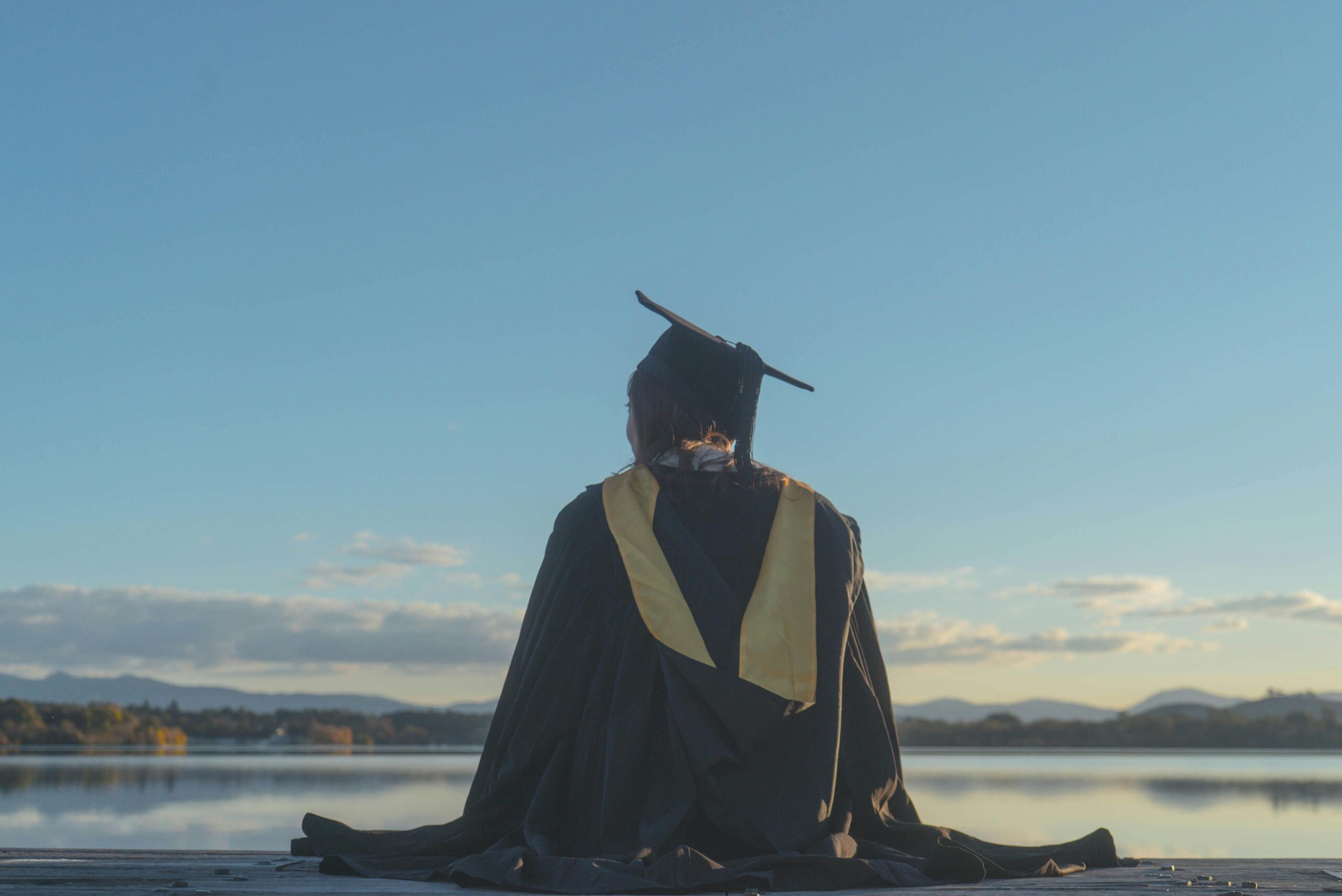 Graduate woman in a cap and gown reflecting by a serene lake at sunset.