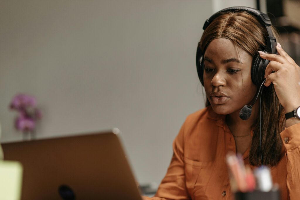 Student Advisory African American woman working in customer support with headphones and laptop indoors.