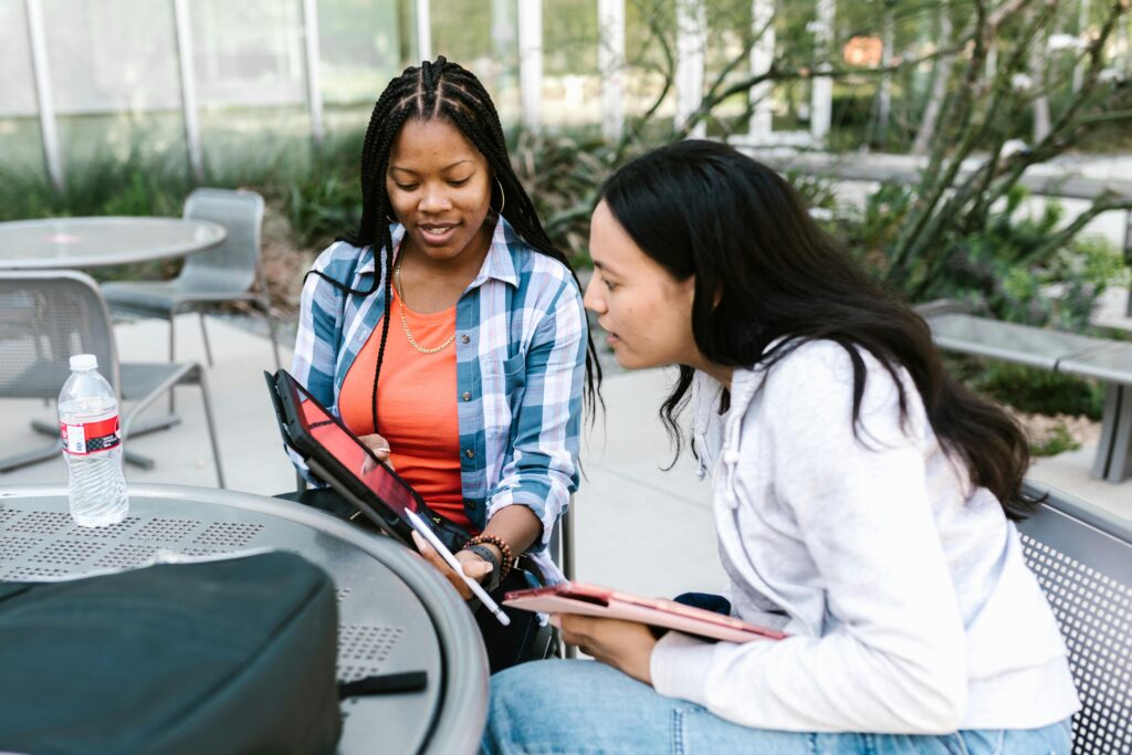 Two young women studying together using a tablet at outdoor campus seating.