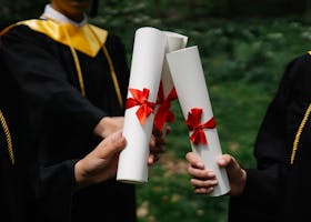 Cost of Attendance Group of graduates holding diplomas with red ribbons during a ceremony outdoors.