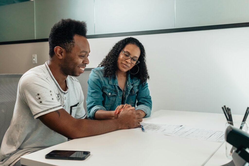 A couple sits at a white table, happily reviewing documents and holding hands in an office setting.